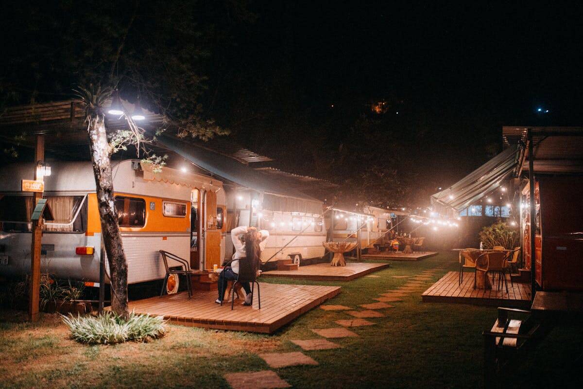 Back view of anonymous female in casual clothes chilling on chair on small terrace near van in trailer park glowing with garlands at night