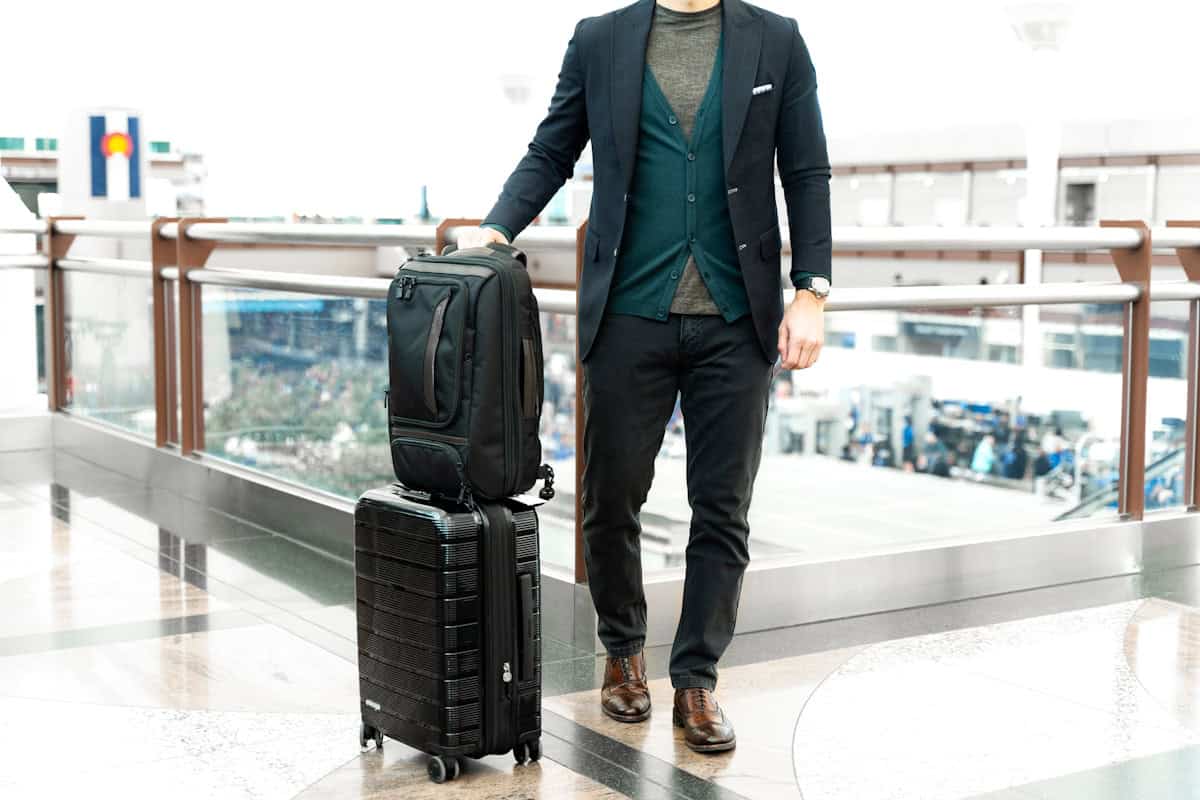 man in blue suit jacket and black pants standing beside black luggage bag